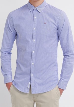 Mężczyzna w dopasowanej, długorękawowej koszuli zapinanej na guziki z pionowymi niebiesko-białymi paskami, beżowe spodnie, ręce w kieszeniach.