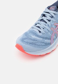 Basket de course bleu clair avec tissu en maille, lacets bleu clair et semelle orange avec détail de logo, sur fond blanc.