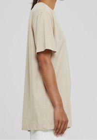 Camiseta de algodón beige de manga corta, corte relajado, escote redondo, con aberturas laterales y textura suave. Mostrada en un modelo de perfil.
