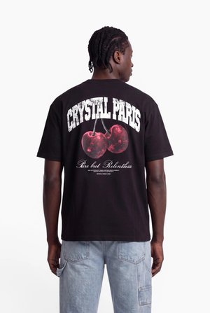 Man with braided hair wearing black t-shirt with "CRYSTAL PARIS" text and red cherries graphic on back, paired with light blue jeans.