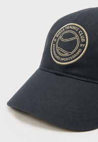 Tmavomodrá baseballová čiapka s prehnutým šiltom a textúrovanou látkou. Na čiapke je kruhová vyšívaná značka v krémovej farbe s tenisovou tematikou.