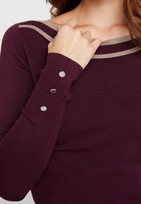 Bordeaux-punainen neule, jossa on ribbitettu pinta, leveä pääntie ja vaaleanbeige akcenttiraitoja. Napit hihoissa tuovat rakenteellista ilmettä.