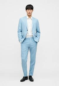 Ανοιχτό μπλε κοστούμι από υφή υφάσματος, με εφαρμοστό σακάκι με δύο κουμπιά, ταιριαστό παντελόνι και λευκό πουκάμισο. Μαύρα επίσημα παπούτσια.