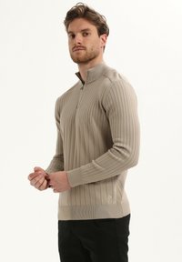 Suéter pullover de manga larga en color beige y de canalé con cuello de cremallera en cuarto y puños ajustados. Presenta un parche con el logo en la parte inferior izquierda.