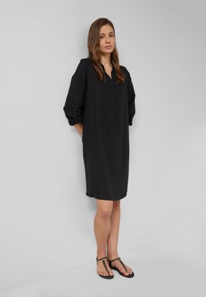 Kobieta stoi przy ścianie, ubrana w luźną czarną sukienkę do kolan z podwiniętymi rękawami i czarne płaskie sandały.