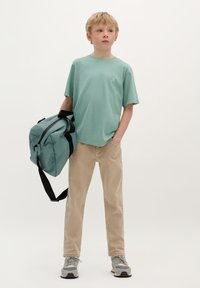 T-shirt vert menthe, pantalon en velours côtelé beige, portant un sac de sport teal avec des sangles noires. Baskets grises. Tenue minimaliste et décontractée.