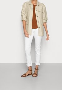 Chaqueta beige de botones con dos bolsillos en el pecho, top marrón, jeans blancos con puños, y sandalias planas marrones con correas en los tobillos. Tejidos suaves.