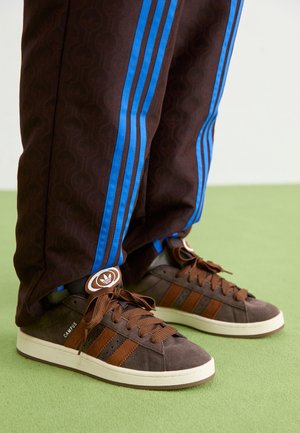 Brązowe sneakersy Adidas Campus z brązowymi sznurowadłami, noszone z brązowymi spodniami z jaskrawoniebieskimi bocznymi paskami, stojące na zielonej podłodze.