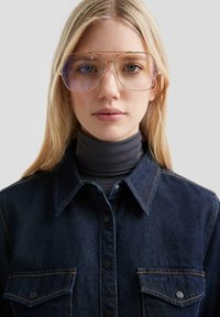 Occhiali da sole rotondi e oversized con montatura dorata e lenti viola chiaro, indossati sopra un dolcevita nero e una camicia in denim scuro con tasche.
