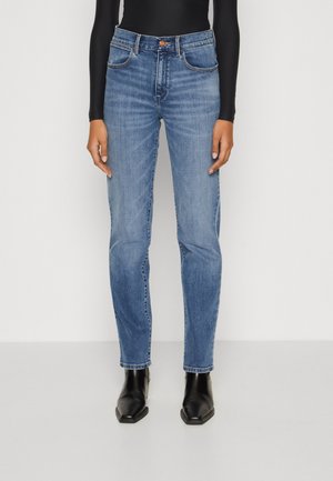 Jean en denim bleu avec une coupe slim, cinq poches, des coutures contrastées et une taille mi-haute. Porté avec des bottines noires à bout pointu.