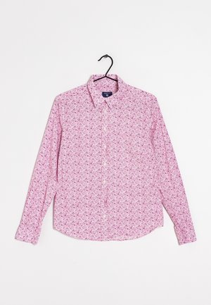 Chemise rose à manches longues avec petits motifs floraux, suspendue sur un cintre noir sur un fond blanc uni.