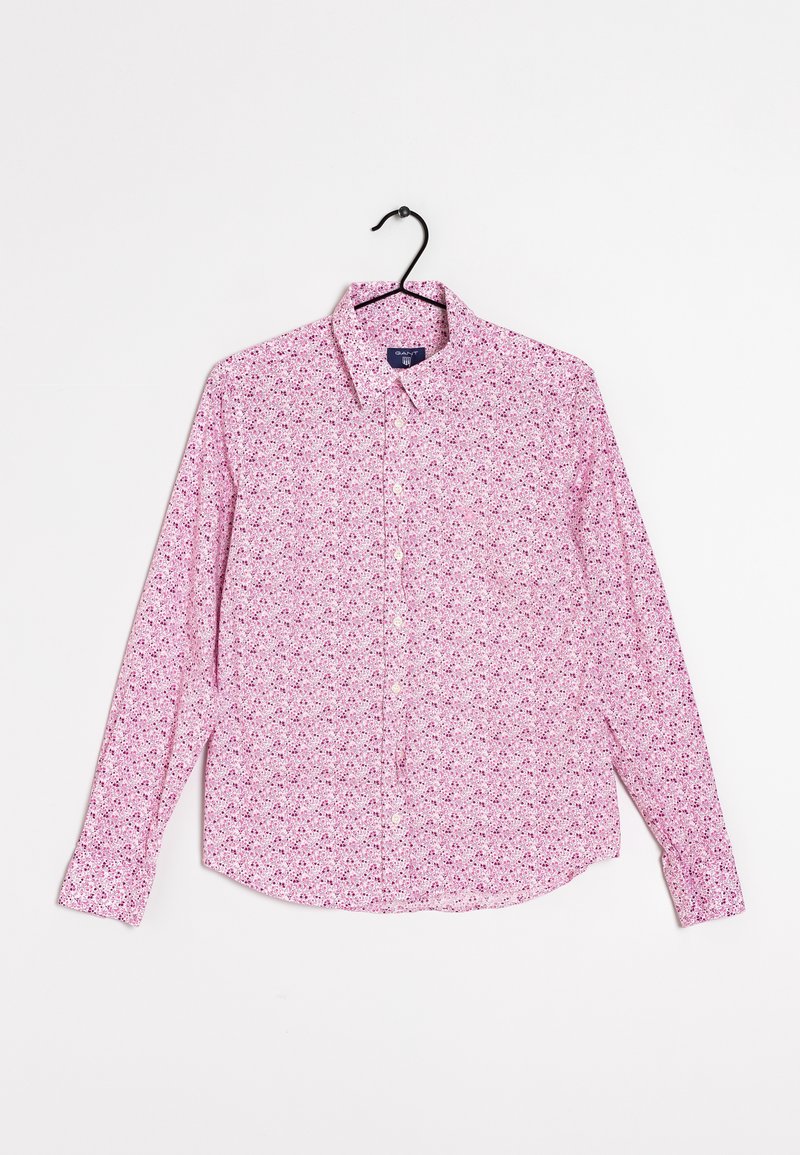 Chemise rose à manches longues avec petits motifs floraux, suspendue sur un cintre noir sur un fond blanc uni.