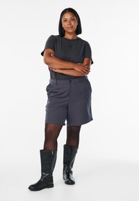 Kvinna står med armarna i kors iförd en mörkgrå t-shirt, grå shorts, svarta leggings och svarta knähöga stövlar på en vit bakgrund.