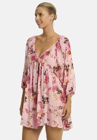 Mujer sonríe mientras lleva un vestido floral rosa con un escote en V profundo y mangas abullonadas, de pie contra un fondo blanco liso.