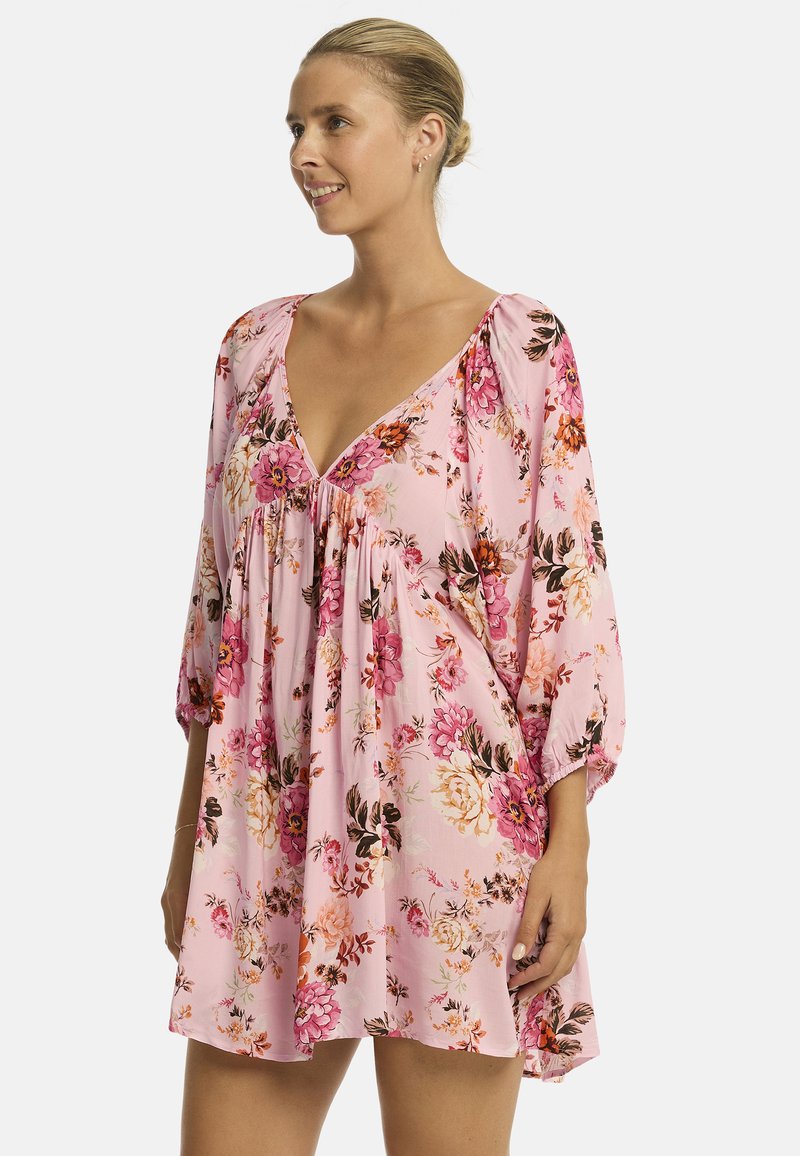 Mujer sonríe mientras lleva un vestido floral rosa con un escote en V profundo y mangas abullonadas, de pie contra un fondo blanco liso.