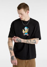 Black short-sleeve t-shirt featuring a colorful graphic of a skeleton holding a drink and food, worn over tan pants.