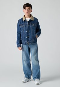 Τζάκετ από τζην με κολάρο από μπεζ χονδρό ύφασμα, διαθέτοντας μπροστινές τσέπες και κούμπωμα με κουμπιά. Συνδυάζεται με φαρδιά μπλε τζιν και λευκά αθλητικά παπούτσια.