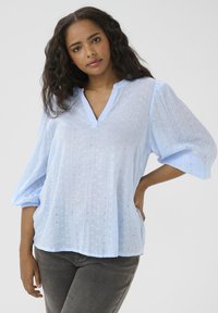 Blouse bleu clair en tissu transparent avec un décolleté en V, des manches bouffantes et de délicats motifs brodés sur toute la surface. Coupe décontractée.