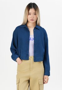 Mladá žena s dlouhými ombre vlasy má na sobě zkrácenou námořnickou košilku s knoflíky přes fialové tričko, spárovanou s béžovými cargo kalhotami, stojí se zahnutýma rukama v kapsách.