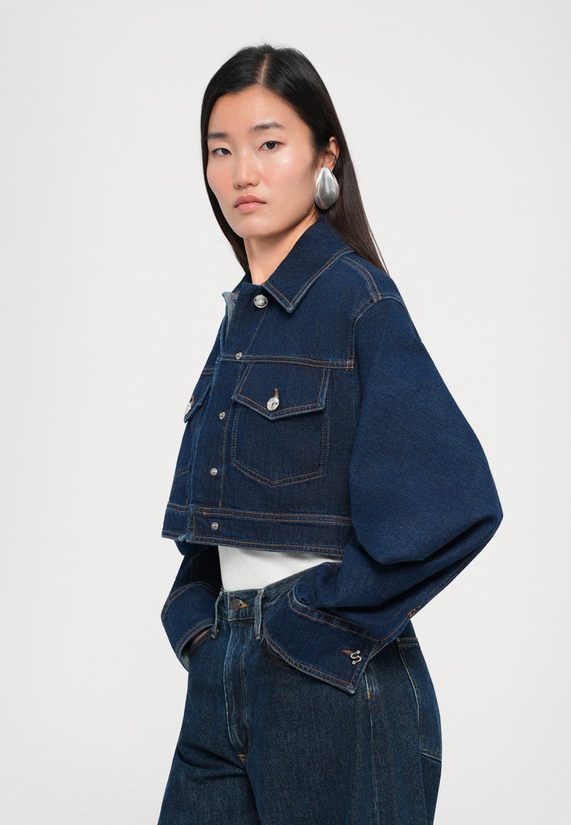 Cropped denim jacket in dark blue, featuring long sleeves, button closure, two chest pockets, and contrast stitching. Worn over a white top.