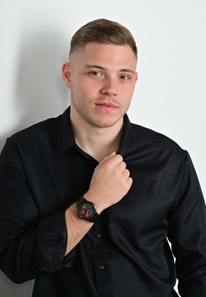 Schwarzes Langarmhemd, das von einer Person getragen wird, mit einer runden schwarzen Uhr mit rotem Design auf dem Zifferblatt, vor einem weißen Hintergrund.