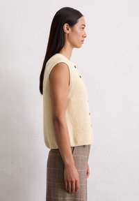 Cremefarbener, gerippter Strickpullover mit kurzen Ärmeln und sechs schwarzen Knöpfen, kombiniert mit braunen Karohosen vor einem neutralen Hintergrund.