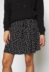 Jupe noire à pois blancs, design plissé, tissu léger. Portée avec un sweat à capuche noir, présentant un style décontracté.