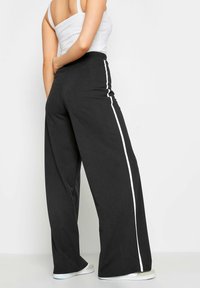 Pantalons noirs à jambes larges en tissu doux, avec des rayures blanches sur les côtés, taille haute et silhouette décontractée.