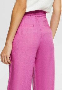 Le bas du torse d'une femme portant un pantalon rose taille haute à jambes larges avec des poches passepoilées à l'arrière et un haut blanc rentré.