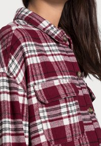 Röd och vit rutiga flanellskjorta med krage, framficka och metallknappar. Mjuk textur med kontrasterande svarta linjer.