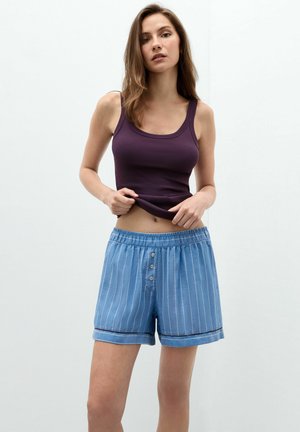 Frau trägt ein dunkelviolettes ärmelloses Oberteil und blaue gestreifte Shorts mit Knöpfen vorne und steht vor einer schlichten hellen Wand.