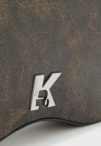 Közelkép egy barna textúrájú bőrfelületről, rajta egy fémes "K" logó, amelyben egy kis kutyasziluett van kivágva.