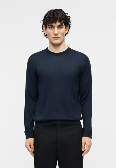 Jeune homme aux cheveux bouclés portant un pull bleu marine uni à manches longues et un pantalon noir, debout devant un fond blanc.