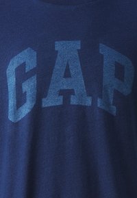 Tmavě modré tričko vyrobené z bavlny, s velkým tučným nápisem "GAP" ve vybledlé modré barvě na přední straně. Lehká textura látky.