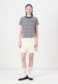 Stribet kortærmet skjorte i navy og hvid, parret med beige shorts med et logo. Modellen har hvide sokker og sorte flade sko på.
