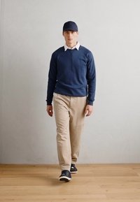 Sudadera navy sobre una camisa blanca con cuello, pantalones beige y una gorra navy. Los zapatos son oscuros con suelas blancas. Modelo caminando sobre un suelo de madera.