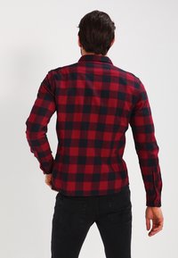 Czerwona i czarna flanelowa koszula w kratę z długimi rękawami, mankietami na guziki i prostym dołem. Materiał wydaje się miękki, z kratkowanym wzorem.