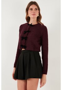 Burgundský zkrácený svetr s texturovanou tkaninou, dlouhými rukávy a třemi černými mašličkovými knoflíky. V kombinaci s černou plisovanou sukní s vysokým pasem.