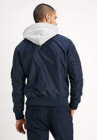 Chaqueta bomber de nailon en navy con capucha gris. Puños y bajo acanalados, detalles acolchados en los hombros y una textura suave y brillante.