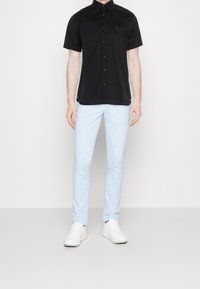 Man in een zwart overhemd met korte mouwen, lichtblauwe slim-fit broek en witte sneakers, staand tegen een effen witte achtergrond.