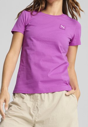 Mujer que lleva una camiseta Puma de color púrpura brillante y pantalones cortos beige claro, con una mano en el bolsillo y cabello castaño ondulado parcialmente visible.