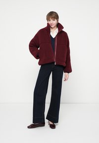 Femeie îmbrăcată cu o jachetă din polar burgundy, o bluză albă, un pulover bleumarin, pantaloni largi închiși la culoare și balerini burgundy, stând în fața unui fundal deschis.