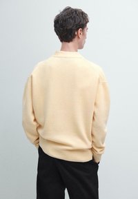 Sv�ětle žlutý svetr s volným střihem, ribbenými manžetami a límcem, zobrazený ze zadní strany. Měkká textura a minimalistický design.