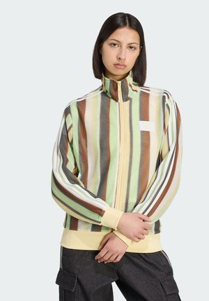 Frau trägt eine mehrfarbige, vertikal gestreifte Jacke mit Reißverschluss und hohem Kragen sowie schwarze Cargohosen, steht mit verschränkten Armen.