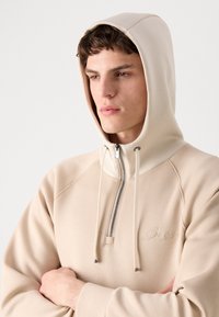 Sweat à capuche beige zippé avec un col haut, des cordons de serrage et un logo brodé. Présente une texture douce et des manches raglan.