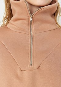 Primer plano de una persona que lleva un sudadera de cuello alto beige con una cremallera plateada en la parte delantera parcialmente subida.