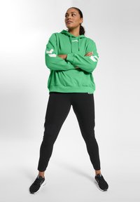 Grön hoodie med en framficka, vita detaljer på ärmarna, kombinerad med svarta leggings och svarta sportskor. Enkelt mönster, avslappnad passform.