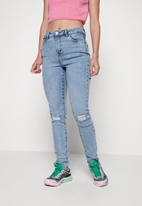 Jeans rasgados azul claro com um corte slim, apresentando um fecho de zíper e botão, emparelhados com ténis verdes e pretos.