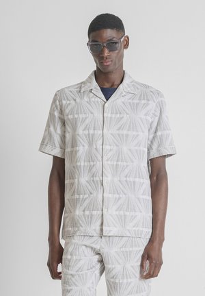 Homme portant une chemise à manches courtes à motif géométrique et un short assorti, accessoirisé de lunettes de soleil rectangulaires foncées.