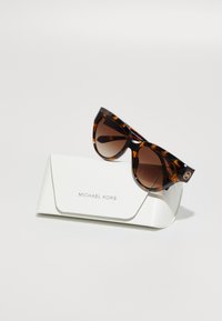 Occhiali da sole cat-eye in tartaruga con lenti sfumate marroni poggiano su un astuccio bianco testurizzato impresso con "MICHAEL KORS."